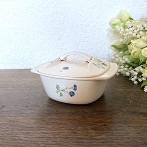 Pode incluir: Travessa de cerâmica creme com tampa e pega. Decorada com padrões florais azuis e verdes. A travessa está sobre uma superfície de madeira.