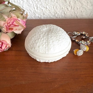 Puede incluir: Una caja de baratijas blanca y redonda con un diseño texturizado similar a un erizo de mar. La caja está sobre una superficie de madera, con algunas joyas y rosas rosas cerca. La caja probablemente se usa para guardar objetos pequeños.