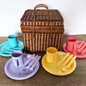 May include: A vintage picnic basket made of woven brown wicker with a handle, surrounded by colorful plastic picnic plates, cups, and cutlery in shades of blue, pink, yellow, and purple. A charming set for outdoor dining.