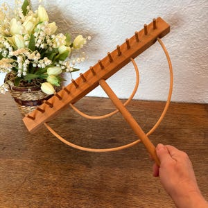 May include: A wooden knitting needle holder with a handle and two circular hoops. The holder has a row of wooden pegs for holding knitting needles.