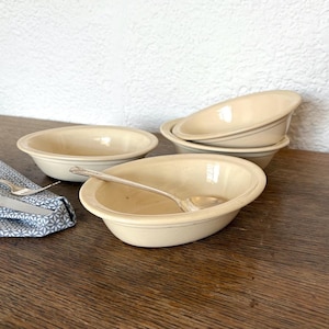 Puede incluir: Conjunto de cuatro cuencos de cerámica de color crema, tres redondos y uno ovalado, con una cuchara de plata descansando en el cuenco ovalado.