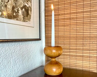 Puede incluir: Un candelero de madera en forma de reloj de arena con una vela blanca encendida. El candelero es de madera marrón claro y tiene un acabado liso y pulido. La llama es de color amarillo y naranja brillante. El candelero está sobre una superficie marrón oscuro.