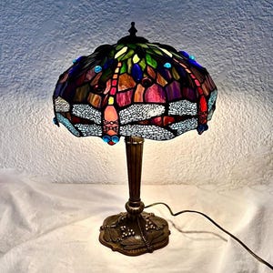 May include: A stained glass table lamp with a dragonfly design. The lamp features a colorful shade with red, blue, green, and purple glass pieces. The base and stem are bronze-colored, and the lamp is plugged into an electrical outlet.
