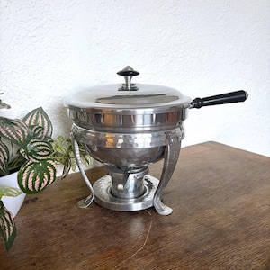 Puede incluir: Un chafing dish plateado vintage con acabado martillado. El plato tiene una tapa con un asa negra y un pomo negro. El chafing dish se asienta sobre un soporte con patas decorativas. El chafing dish está sobre una superficie de madera.