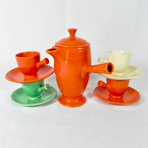 May include: A set of four vintage ceramic coffee cups and a matching teapot in orange, green, and white. The cups have a unique spout design and are stacked on their saucers.