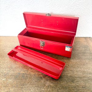 May include: A bright red metal tool box with a hinged lid and a single metal latch. The box includes a removable red metal tray. A small rectangular sticker with the text Mastercraft and a maple leaf logo is visible on the front right side.