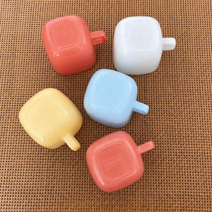 Peut inclure: Un ensemble de cinq tasses carrées aux couleurs corail, blanc, bleu clair et jaune. Chaque tasse possède une anse latérale et le logo Glasbake moulé sur le fond. Les tasses sont disposées en groupe, révélant leur finition lisse et brillante.
