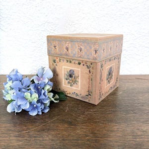 Könnte beinhalten: Eine quadratische, dekorative Box mit Deckel, mit floralen und botanischen Designs. Die Box hat einen hellbraunen Hintergrund mit quadratischen Blumenbildern und einem blau-goldenen Muster. Ein Strauß blauer Hortensien liegt neben der Box.