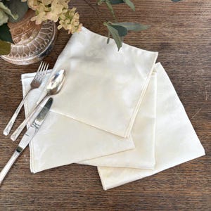 May include: Three cream-coloured cloth napkins with a delicate floral pattern are stacked on a wooden surface. Silverware, including a fork, spoon, and knife, is arranged to the left of the napkins. Decorative flowers are in the background.