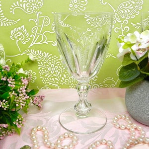 May include: A clear crystal wine glass with a decorative stem and etched floral design. The glass has a wide bowl and sits on a round base. The background features a green floral pattern and pink flowers.