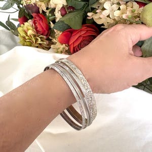 Puede incluir: Un conjunto de pulseras plateadas con intrincados diseños florales. Las pulseras están apiladas en una muñeca, mostrando su diseño detallado. Las pulseras son de color plata clara, y los diseños florales están grabados en el metal.