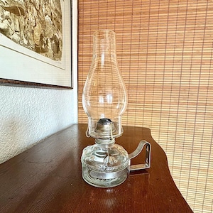 Puede incluir: Una lámpara de aceite de vidrio transparente con asa y base de metal. La lámpara tiene una gran chimenea de vidrio redondeada y un depósito de vidrio grabado. La lámpara está sobre una superficie de madera oscura.