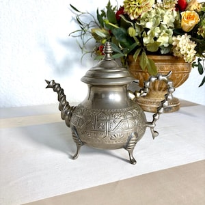 May include: A silver teapot with detailed embossed designs and a twisted spout. The teapot features a lid with a finial and rests on three small feet. The handle is a decorative loop. A vintage tea accessory.