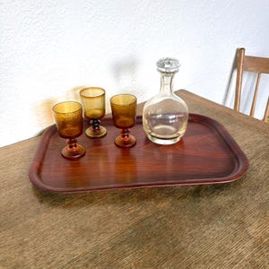 May include: A vintage serving set comprising a rectangular wooden tray, three amber-tinted stemmed glasses, and a clear glass decanter with a stopper. The glasses and decanter are arranged on the tray, which is placed on a wooden table.
