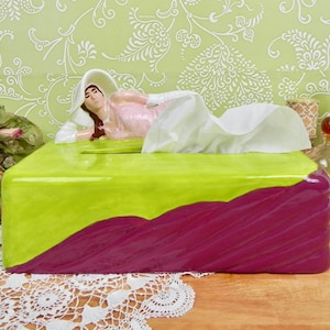 May include: A green and purple ceramic tissue box cover with a figurine of a woman lying on top. The woman is wearing a white dress and has long brown hair.