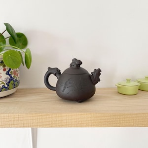 Peut inclure: Une théière en céramique brun foncé avec une petite figurine d'animal sur le couvercle. La théière a un corps rond et un bec verseur sur le côté. La théière est posée sur une étagère en bois.