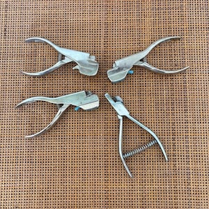 May include: Four silver metal tools with curved handles and spring mechanisms. The tools have a scissor-like design, with a blue accent on the top. They are arranged on a brown woven surface.