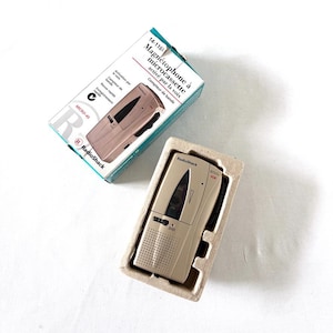 May include: A beige RadioShack microcassette recorder with a speaker grille, control switches, and VOX label, housed in a molded cardboard tray. Beside it is the original product box featuring the text RadioShack and Magnétophone à microcassette.