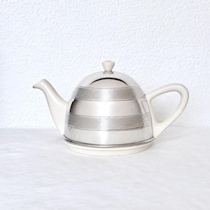 May include: A teapot featuring a polished silver-tone body with two horizontal bands of textured cross-hatch detailing. The teapot includes a matching cream-coloured handle, spout, base, and a small knob on the lid, creating a contrast between the metallic finish and the solid cream components.