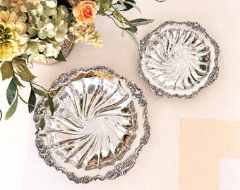 Könnte beinhalten: Zwei runde Servierschalen aus silberfarbenem Metall mit einer polierten, spiralförmig geriffelten Mitte und einem dekorativen, geprägten Blumenrand. Die Schalen sind neben einem Strauß künstlicher Blumen in Rosa-, Gelb- und Grüntönen angeordnet.