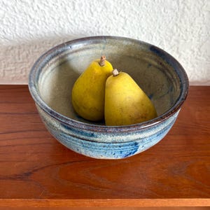 May include: A handcrafted ceramic bowl with a blue and grey glaze contains two ripe yellow pears. The bowl is wide and shallow, resting on a wooden surface. The pears have brown stalks and are a vibrant yellow colour.