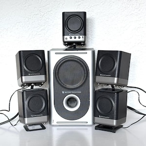 Könnte beinhalten: Ein silbernes und schwarzes Altec Lansing Computer-Lautsprechersystem. Das System umfasst einen zentralen Subwoofer und fünf Satellitenlautsprecher. Jeder Lautsprecher hat ein schwarzes Mesh-Gitter und einen silbernen Sockel. Kabel gehen von den Lautsprechern ab.