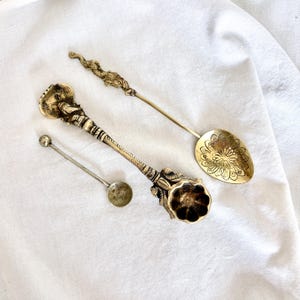 Könnte beinhalten: Drei Messinglöffel in verschiedenen Größen und Ausführungen. Ein Löffel hat einen langen Stiel mit Drachenmotiv und eine blumengravierte Laffe. Ein weiterer hat einen verzierten Stiel mit einer blütenförmigen Laffe. Der dritte ist ein kleiner, schlichter Löffel.