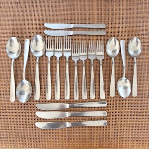 以下が含まれることがあります： 織られた茶色の表面に配置された、スプーン、フォーク、ナイフを含むステンレス鋼のカトラリーのコレクション。カトラリーは、光を反射するシンプルでエレガントなデザインです。配置はフォーマルなダイニング環境を示唆しています。