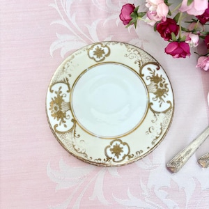 Może przedstawiać: Stos antycznych, kremowych spodków ze złotymi wzorami kwiatowymi i zwojami. Spodki mają złote obramowanie i biały środek. Talerze leżą na różowym obrusie z kwiatowym wzorem.