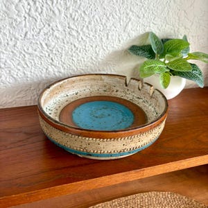 Puede incluir: Un cenicero redondo de cerámica con un centro turquesa, rodeado por un anillo marrón y una banda beige con una raya azul. El cenicero tiene cuatro muescas para cigarrillos.