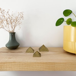 May include: Three gold-coloured metal pyramid-shaped decorative objects of varying sizes on a wooden shelf.