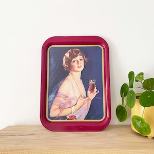 May include: A vintage burgundy serving tray with a Coca-Cola advertisement featuring a woman in a pink dress holding a glass of Coca-Cola. The tray has a rectangular shape with rounded corners and a gold border.