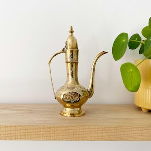 May include: A decorative brass pitcher with an intricate floral design. The pitcher features a long, curved spout, a handle, and a lid. The body is adorned with etched patterns. It is displayed on a wooden shelf, next to a plant in a yellow pot.