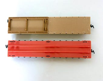 May include: Overhead view of two model train flatcars. One is tan with a wooden deck design, the other is red. Both have black couplers and are set against a white background. These are likely for a model train set.