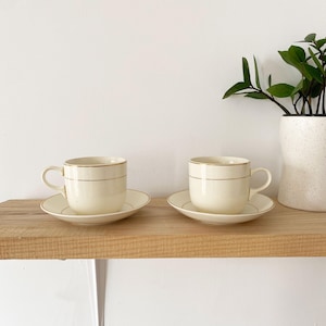Peut inclure: Deux tasses à thé en céramique blanche avec une bordure dorée, chacune sur une soucoupe assortie, posées sur une étagère en bois.