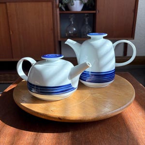 Könnte beinhalten: Zwei weiße Keramik-Teekannen mit blauen Streifen. Eine Teekanne ist rund, die andere höher. Beide haben passende blaue Deckel und Griffe. Sie stehen auf einem Holztablett, mit einem Holzschrank im Hintergrund.