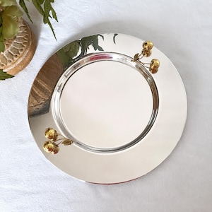 May include: A round, polished silver-tone metal serving tray featuring a smooth, reflective surface. Two decorative gold-tone cherry motifs, each consisting of two small spheres and a leaf, are attached to the rim on opposite sides of the tray.