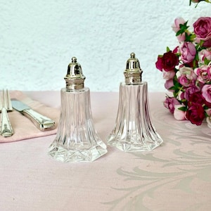 Könnte beinhalten: Zwei Salz- und Pfefferstreuer aus klarem Glas mit silberfarbenen Deckeln. Die Streuer haben ein ausgestelltes, geripptes Design. Links sind eine Gabel und ein Messer zu sehen, rechts rosa Blumen. Auf einer rosa Oberfläche.