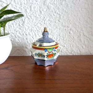 Puede incluir: Un azucarero pequeño de cerámica vintage con tapa. El cuenco es azul claro con un diseño geométrico y detalles florales en naranja, amarillo y verde. La tapa está rematada con un adorno dorado. El cuenco está sobre una superficie de madera.