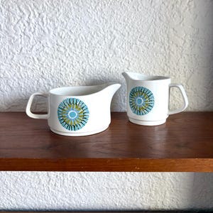 Puede incluir: Dos jarras de crema de cerámica blanca con un diseño circular azul, verde y amarillo. Las jarras tienen un asa y un pico. Están sobre una estantería de madera marrón oscuro contra una pared blanca texturizada.