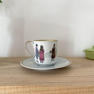 May include: A white porcelain teacup and saucer with a gold rim. The cup features a colorful illustration of people in traditional clothing.