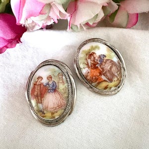 Peut inclure: Deux boucles d'oreilles ovales vintage avec des montures en métal argenté. Chaque boucle d'oreille présente une scène miniature peinte à la main de personnages en costumes d'époque. L'arrière-plan comprend des arbres et un ciel clair. Les boucles d'oreilles sont posées sur un tissu blanc.