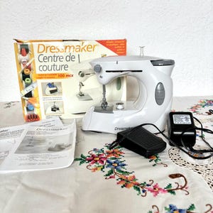 May include: A white Dressmaker sewing machine with a black foot pedal and power adapter. The machine is next to a box with the text "Dressmaker Centre de couture". The machine sits on a floral embroidered tablecloth.