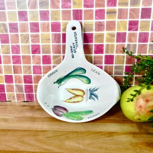 Puede incluir: Separador de salsa blanco de cerámica con asa. El separador tiene un diseño de verduras verdes, amarillas y moradas y las palabras "No-Drip Gravy Separator", "Gravy", "Lean" y "Gravy" impresas en él.