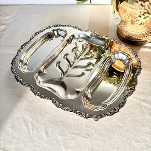 May include: A silver-plated serving tray with three compartments and ornate handles. The tray has a decorative, scalloped edge and a central leaf-like design. The reflective surface of the tray catches the light, highlighting its intricate details.