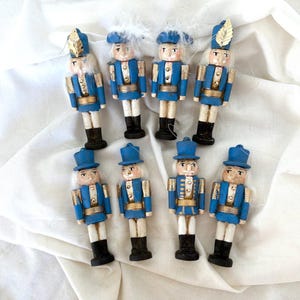 May include: Eight miniature wooden nutcracker figurines. Each is painted with a blue jacket, gold accents, and black boots. Some have white hair and others have a blue hat with a gold leaf.