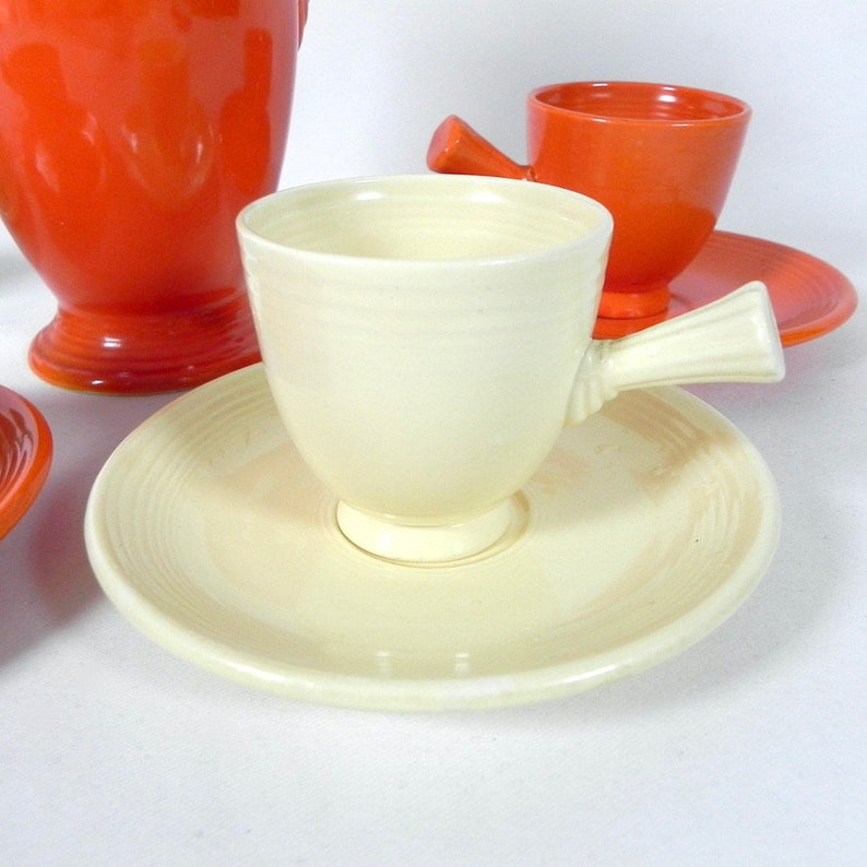 May include: A set of vintage teacups and saucers. The teacups are white with a unique handle and the saucers have a subtle pattern. The teacups are paired with orange saucers.