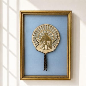 Op de afbeelding: Een ingelijste decoratieve handwaaier met een centraal goudkleurig adelaarembleem met gespreide vleugels, omringd door een radiaal patroon van dunne metalen spaken en kleine blauwe accenten, op een lichtblauwe stoffen achtergrond in een gouden lijst.
