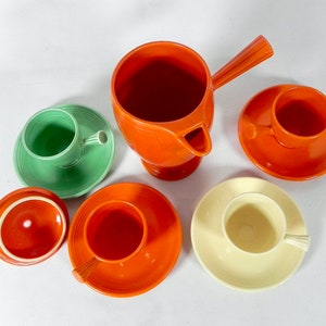 May include: A set of orange, green, and cream colored ceramic teacups and saucers. The set includes a teapot with a handle and a spout.