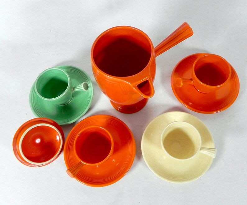 May include: A set of orange, green, and cream colored ceramic teacups and saucers. The set includes a teapot with a handle and a spout.